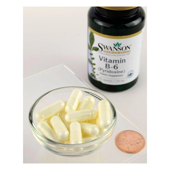 A small glass bowl with white capsules is next to a bottle of Swanson Vitamin B6 Pyridoxine 100 mg 100 Capsules, highlighting its importance for energy metabolism and cardiovascular health, with a penny for scale.