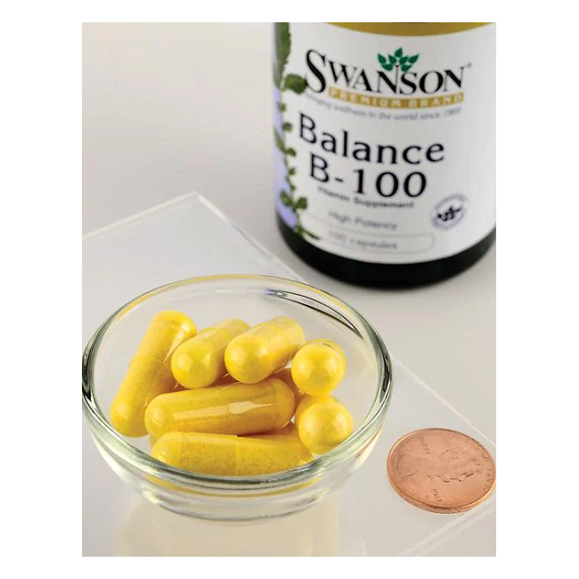 A glass bowl with yellow capsules sits beside a penny, with a Swanson Balance Vitamin B-100 Complex 100 Capsules bottle in the background, emphasizing its essential B vitamins for metabolic support.