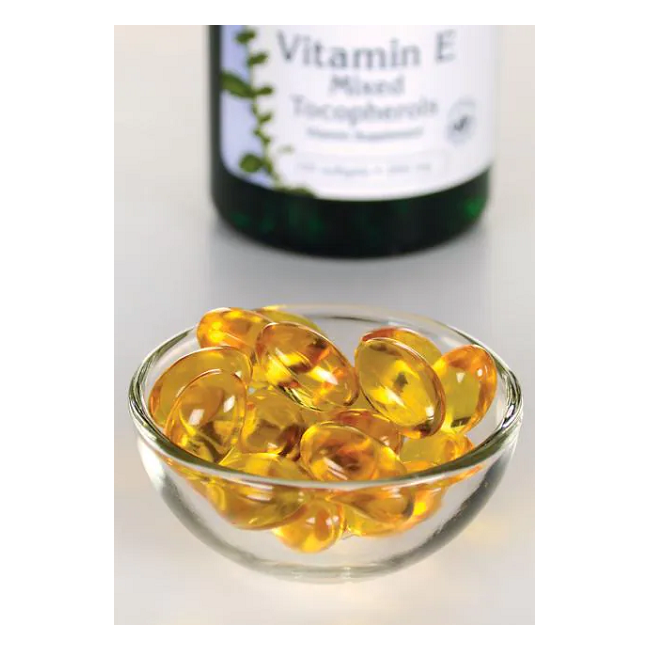 A small glass bowl contains yellow gel capsules, while a Swanson Vitamin E - 400 IU 100 softgel Mixed Tocopherols bottle is blurry in the background, promoting antioxidant support for cardiovascular health.