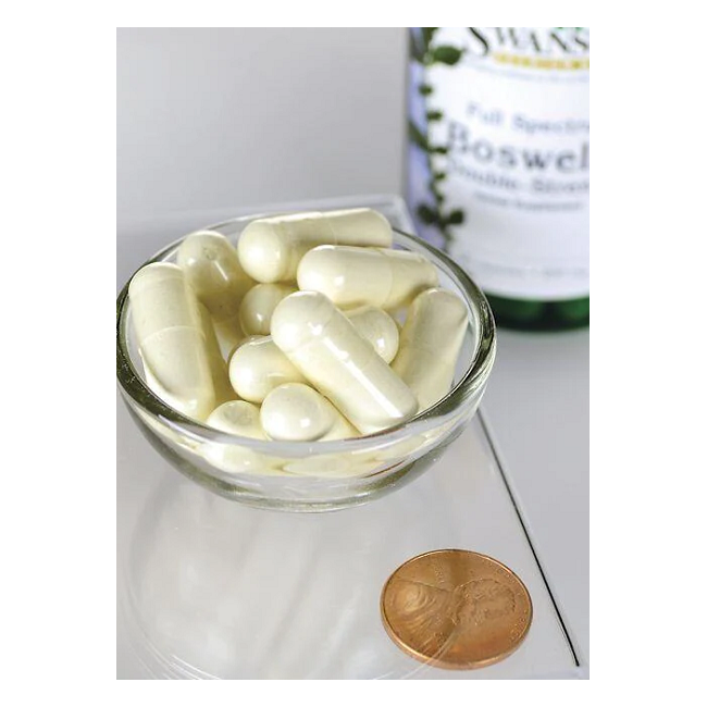 A small glass bowl filled with white capsules sits on a reflective surface beside a penny, with a Swanson Boswellia 800 mg 60 Capsules bottle, known for joint health benefits in Ayurvedic tradition, in the background.