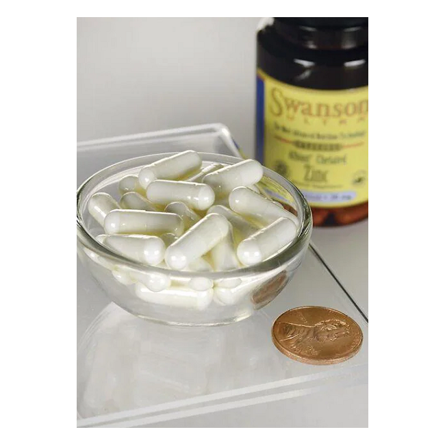 A small glass bowl with white capsules lies beside a "Swanson" bottle labeled "Zinc - 30 mg 90 capsules Albion Chelated," featuring metabolism support supplements, with a penny for scale.