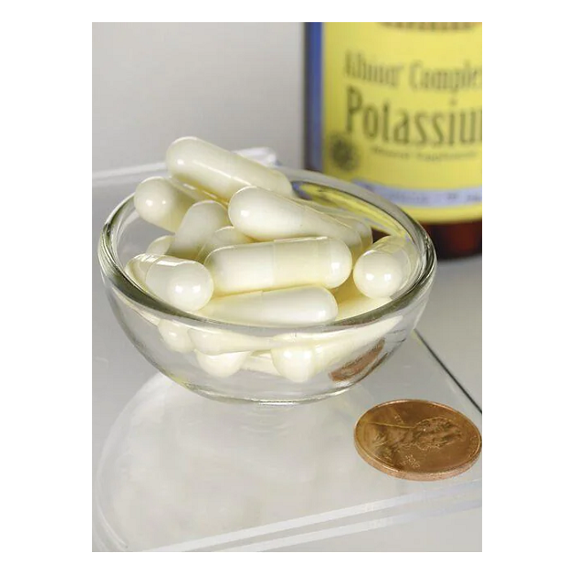 A small glass bowl filled with white capsules is shown next to a penny for scale. In the background, a blurred bottle of Swanson's Albion Potassium 99 mg 90 Capsules promises electrolyte balance support.