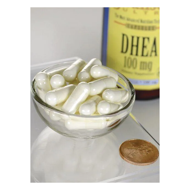 A small glass bowl holds white capsules next to a Swanson bottle labeled "DHEA 100 mg 60 Capsules - Maximum Strength," often called the youth hormone, with a penny for scale.