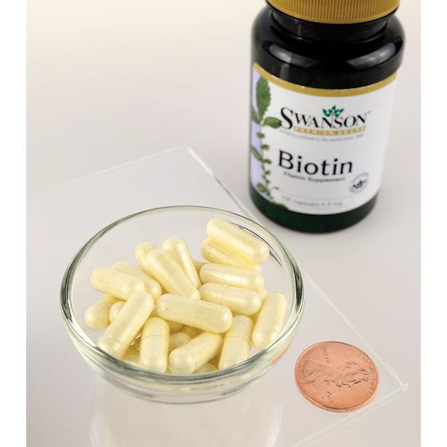 Um frasco de suplemento alimentar Swanson Biotin - 5 mg 100 capsules ao lado de uma moeda sobre uma mesa.
